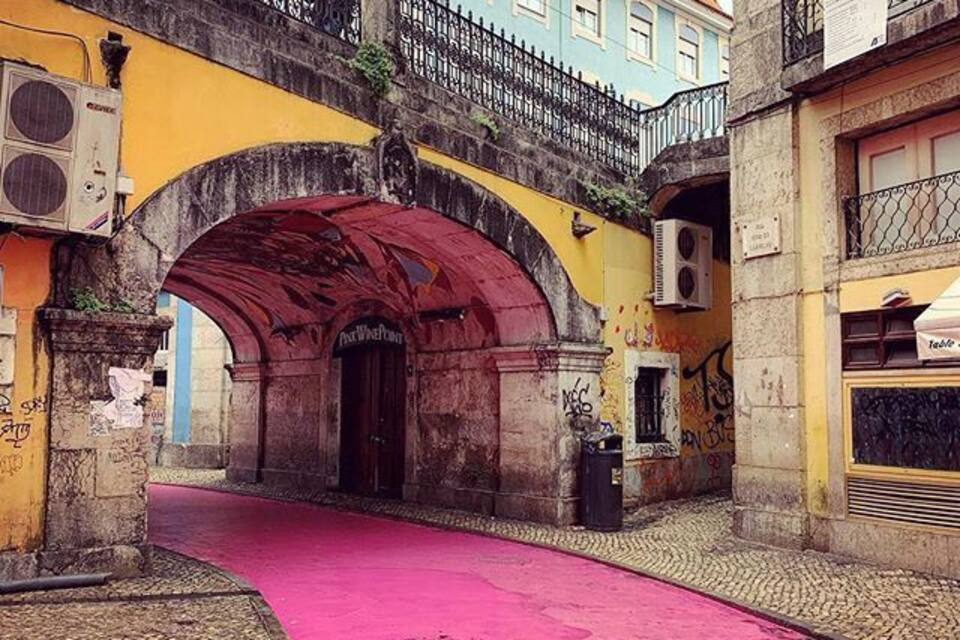 A street with a pink pavement, an old stone archway, and a building with a blue facade.