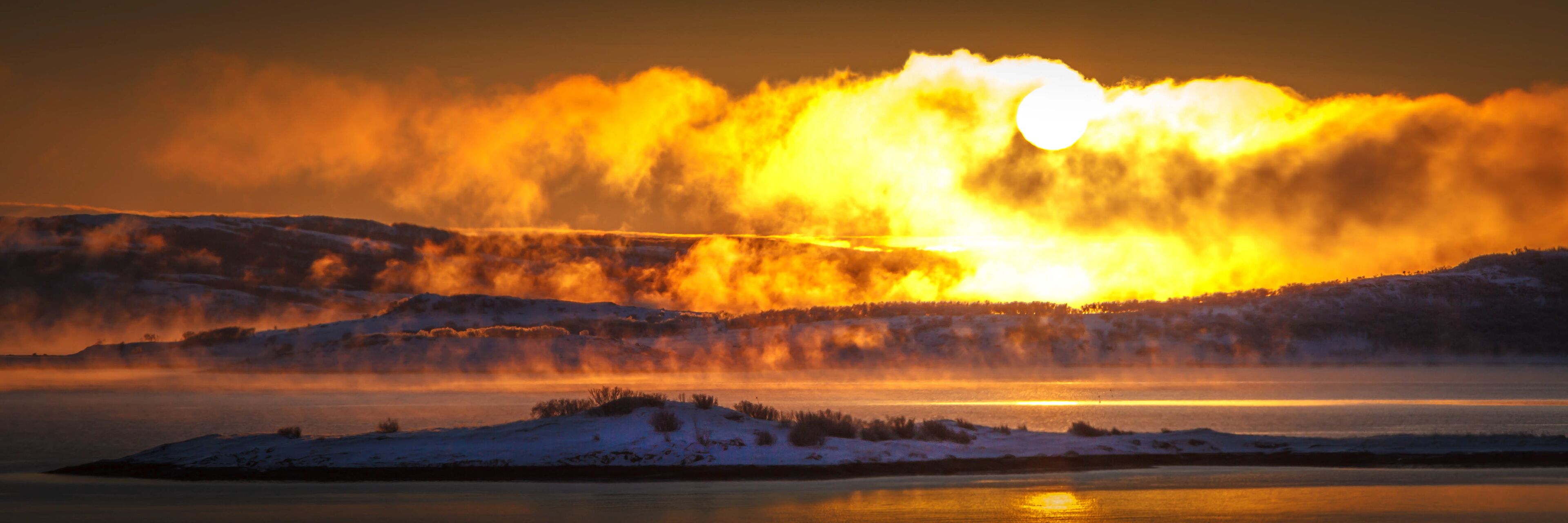 A sunset over a snow-covered landscape with a body of water in the foreground.