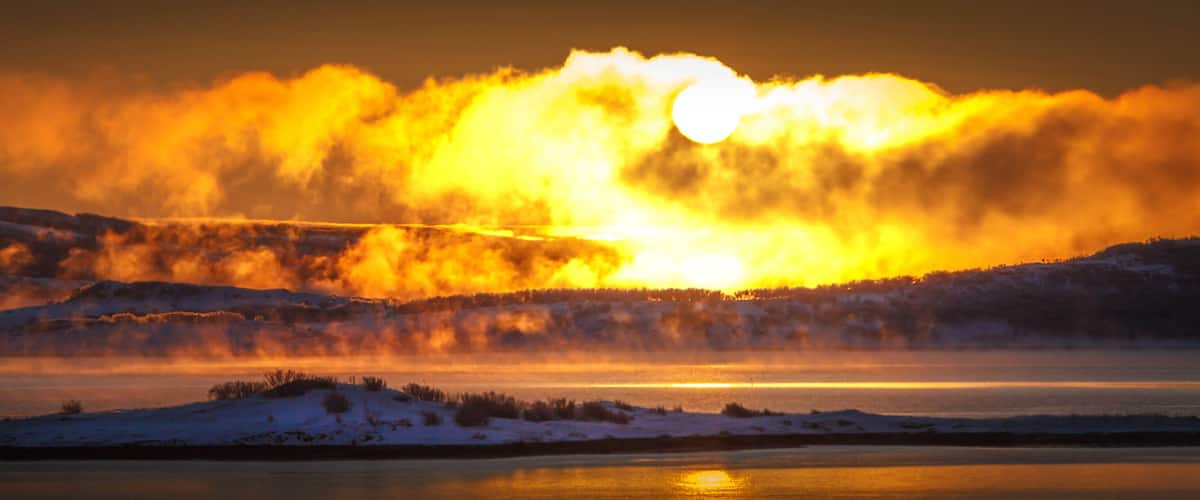 A sunset over a snow-covered landscape with a body of water in the foreground.