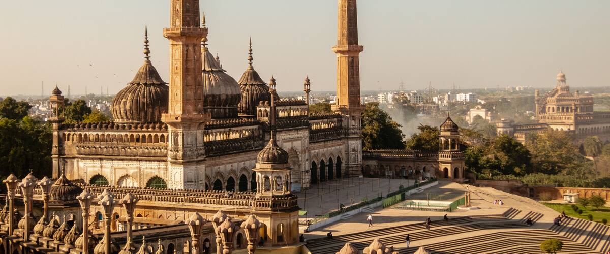 A historic building with multiple domes and minarets, surrounded by a large open courtyard with patterned paving.