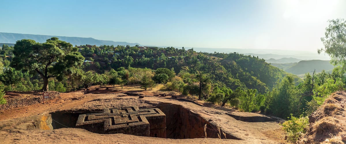 A large ancient stone structure with carvings, surrounded by a forested hillside and distant mountains.