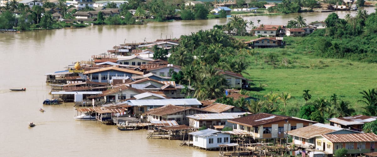 A riverside settlement with houses built on stilts, surrounded by lush greenery and hills in the background.