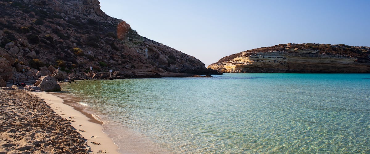 A clear turquoise sea, sandy beach, rocky cliffs, and a clear sky.