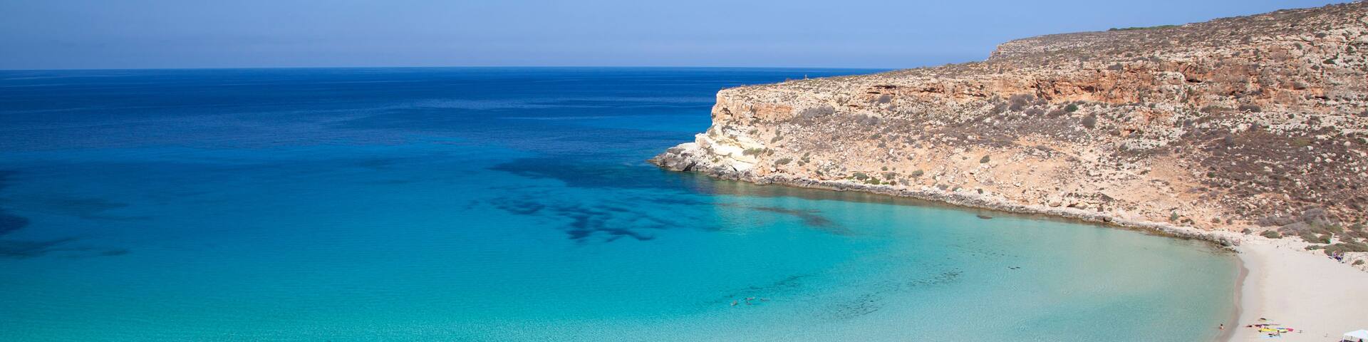 A coastal landscape with clear turquoise waters, a sandy beach, and rocky cliffs.