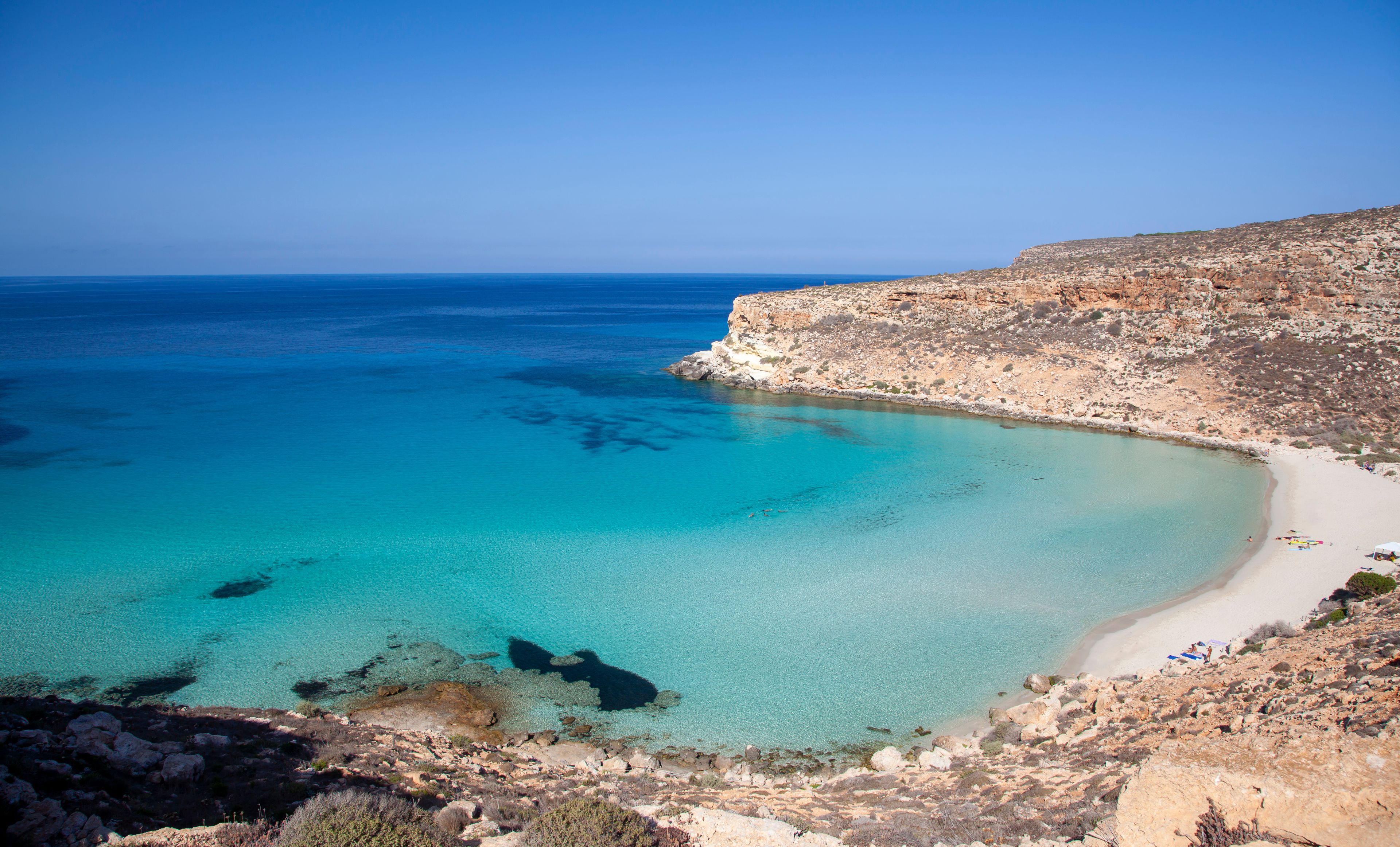 A coastal landscape with clear turquoise waters, a sandy beach, and rocky cliffs.