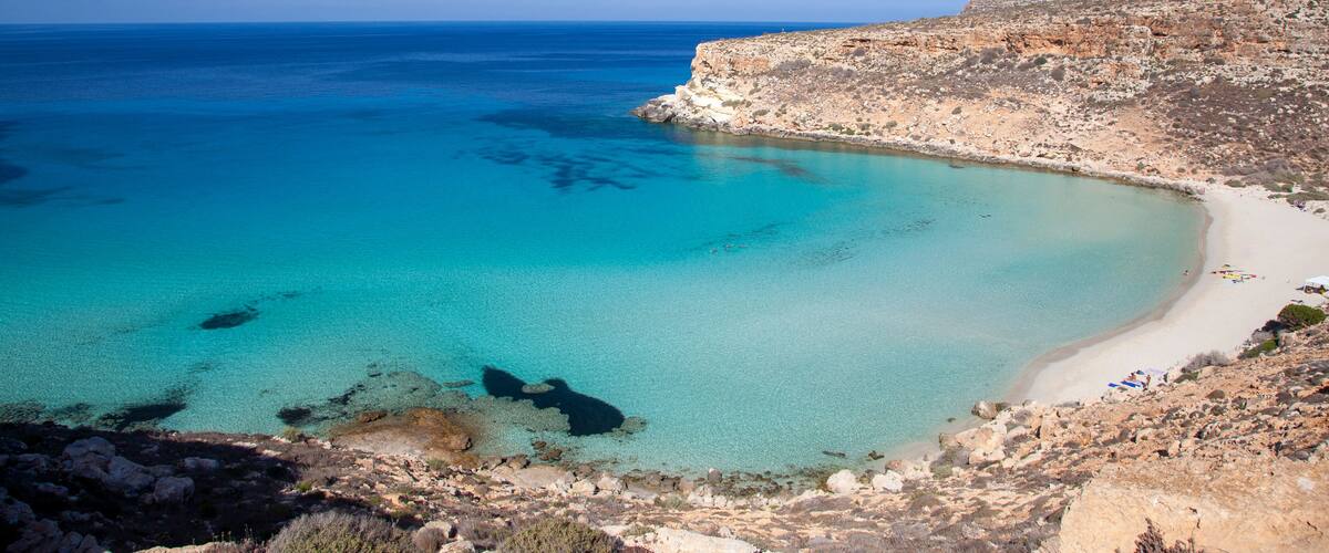 A coastal landscape with clear turquoise waters, a sandy beach, and rocky cliffs.