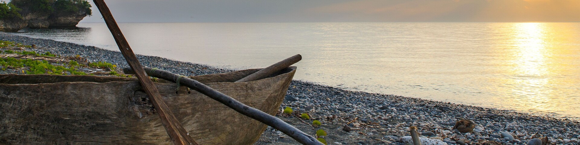 A wooden boat on a pebbly beach with a sunset in the background.
