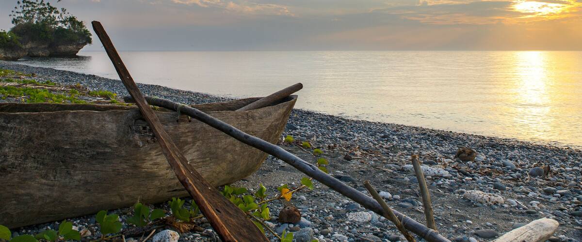 A wooden boat on a pebbly beach with a sunset in the background.