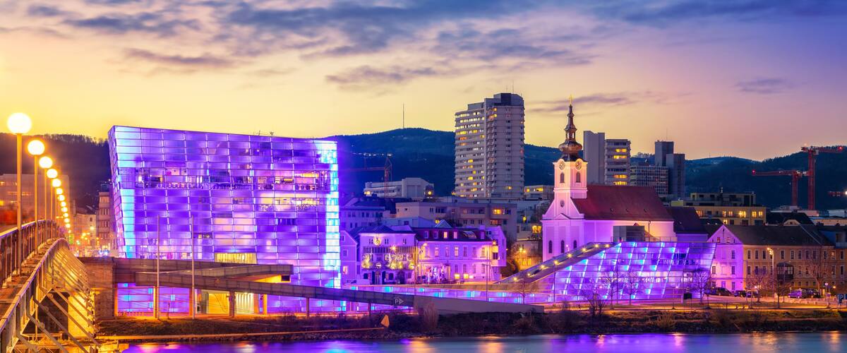 A cityscape at dusk with illuminated buildings, a bridge, and a river reflecting the lights.