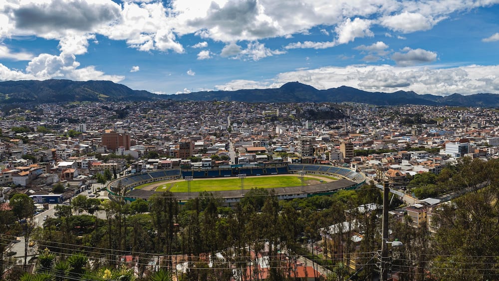 Eine Stadtlandschaft mit einem großen Stadion, umgeben von Wohngebäuden und Bergen im Hintergrund.