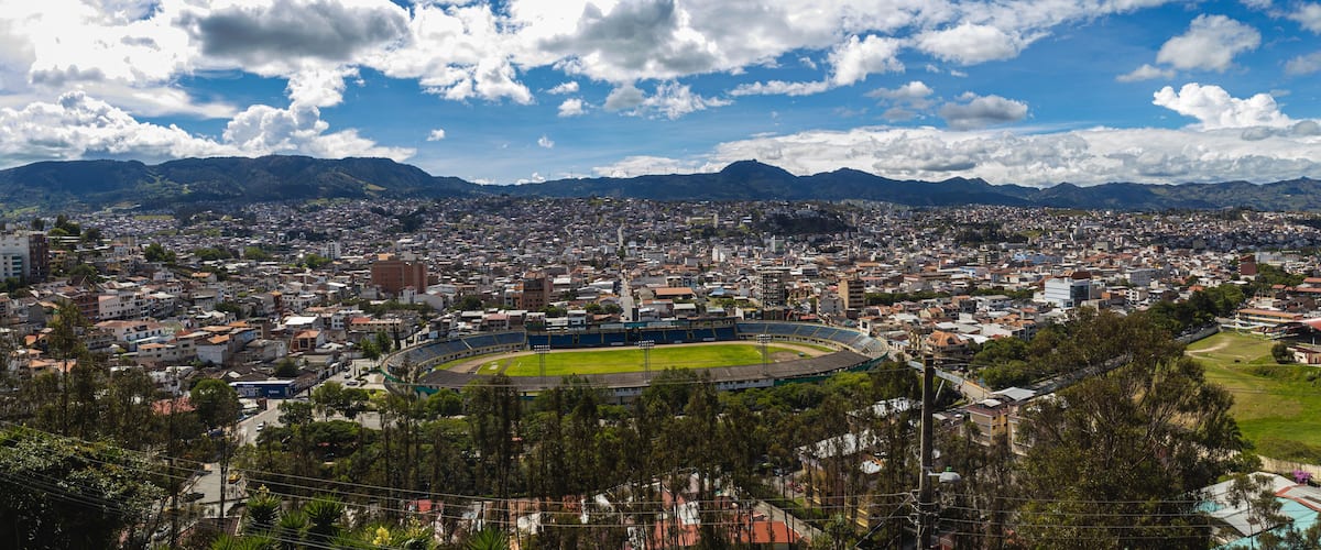 Eine Stadtlandschaft mit einem großen Stadion, umgeben von Wohngebäuden und Bergen im Hintergrund.