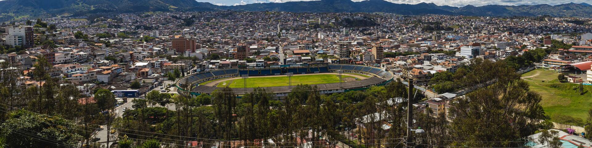 A cityscape with a large stadium surrounded by residential buildings and mountains in the background.