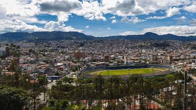 Un paisaje urbano con un estadio grande rodeado de edificios residenciales y montañas al fondo.