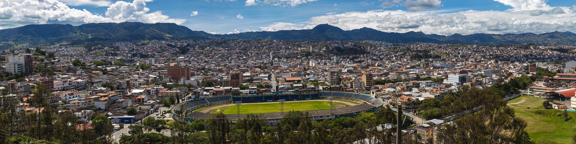 Uma paisagem urbana com um estádio grande, cercado por edifícios residenciais e montanhas ao fundo.