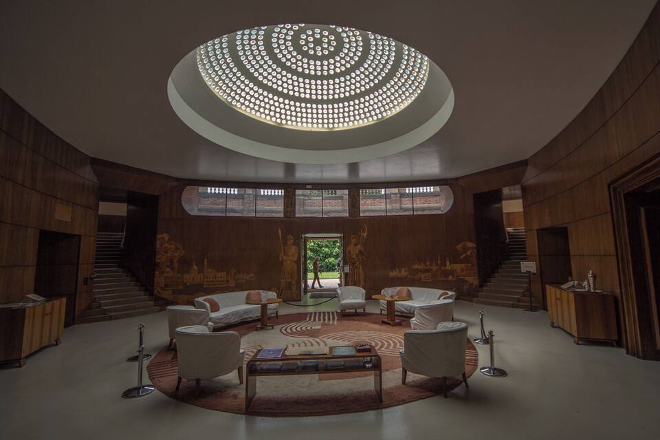 Un intérieur spacieux avec un luminaire circulaire au plafond, un espace salon central et un escalier menant à une zone extérieure.