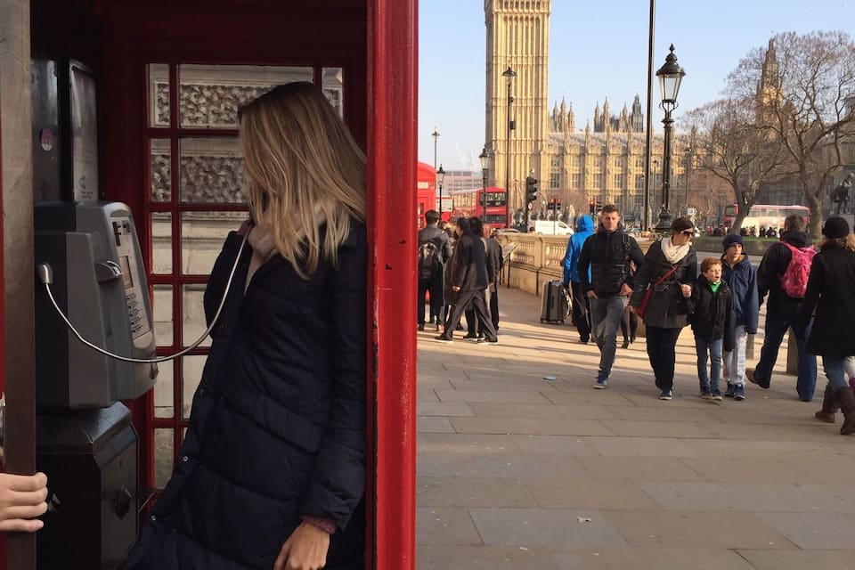Uma cabine telefônica vermelha com uma pessoa entrando, uma torre do relógio ao fundo e pessoas caminhando na rua.