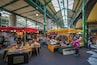 A bustling indoor market with various stalls, including a cheese stand and a produce market.
