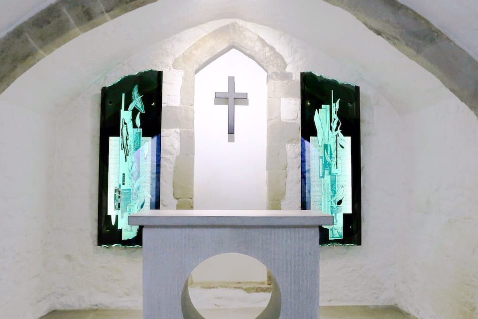 Uma capela vazia com uma cruz, uma parede de pedra e janelas com vitrais.