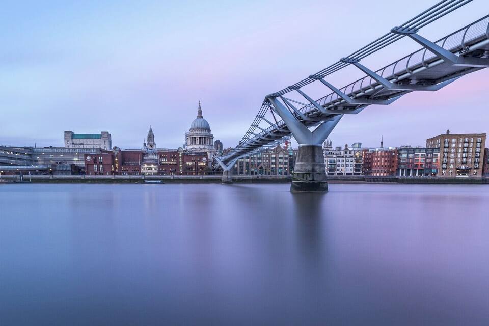 A modern bridge over a calm river with historic buildings in the background.