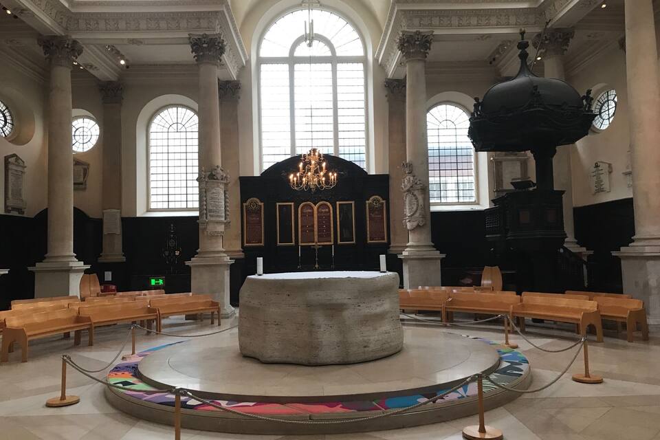 Un salón imponente con techos altos, amplias ventanas en arco y un altar de piedra central rodeado de bancas de madera.