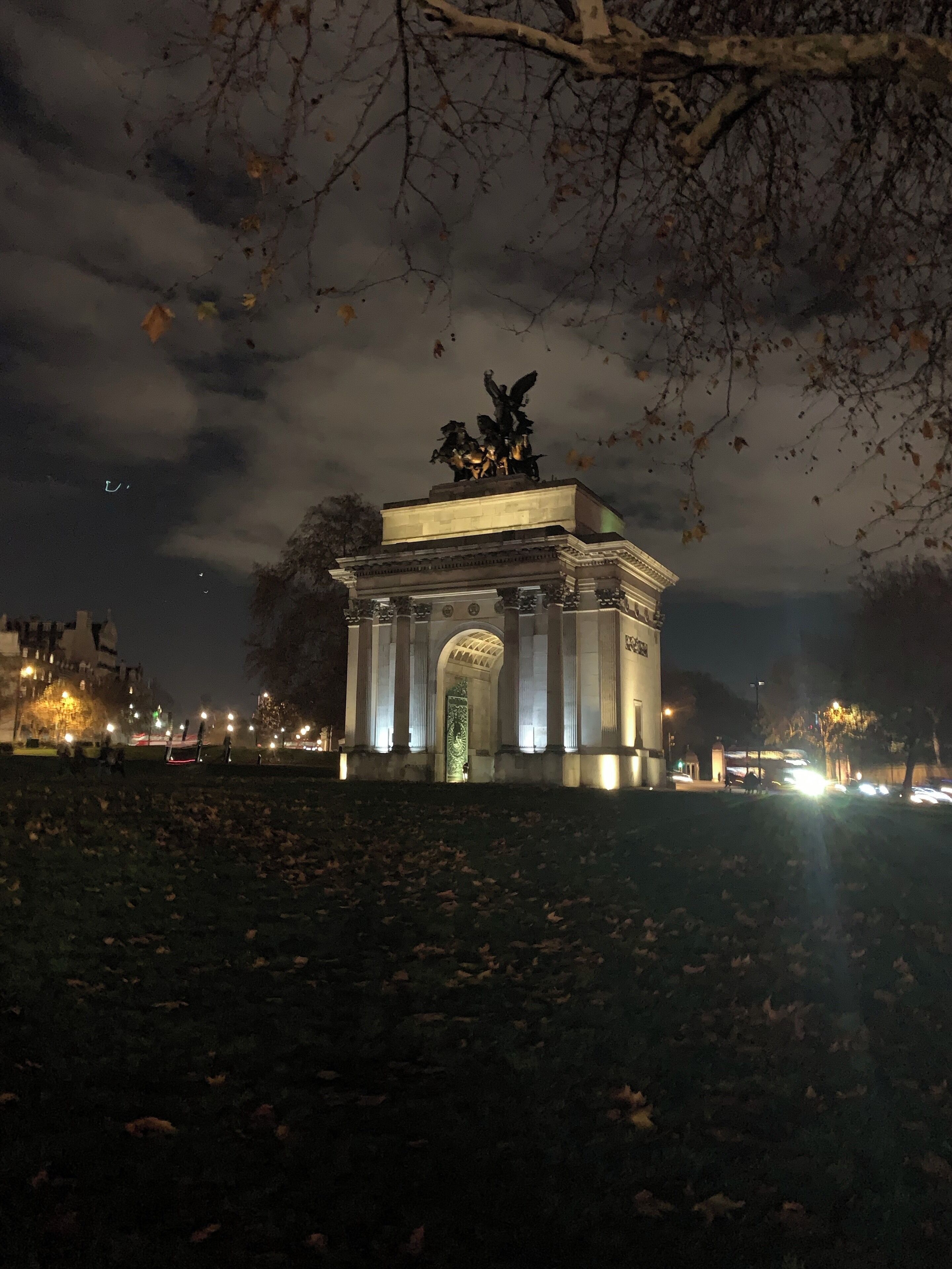 A nighttime view of a classical monument with a statue on top, surrounded by trees and illuminated by streetlights.