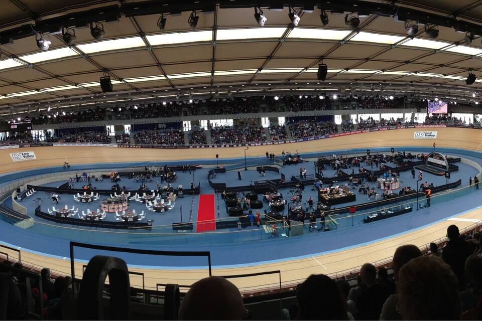 An indoor velodrome with a blue track, surrounded by spectator stands.