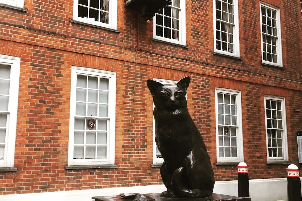 Uma estátua de gato preto sobre um pedestal, em frente a um edifício de tijolos vermelhos com janelas brancas e um relógio acima.