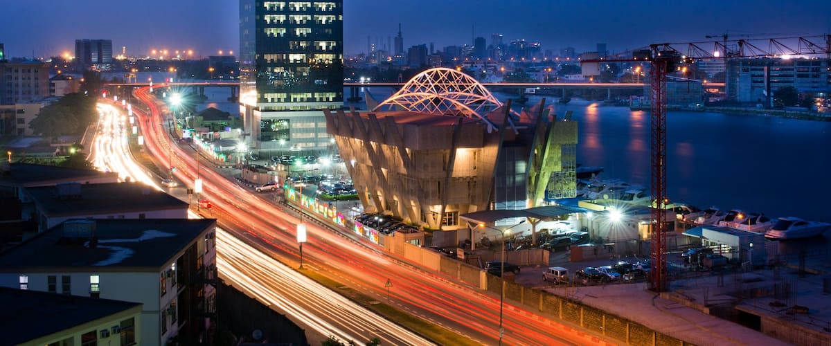 A cityscape at dusk with illuminated buildings, a prominent tower, and a bridge with light trails.