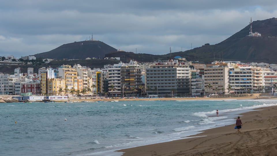 A coastal cityscape with a beach, buildings, and mountains in the background.