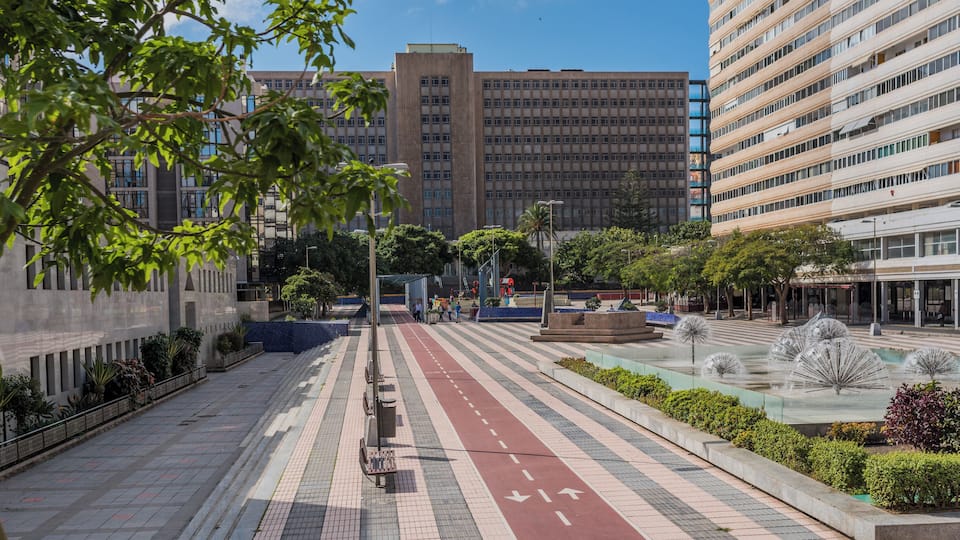 A city street with a pedestrian walkway, a fountain, and modern buildings.