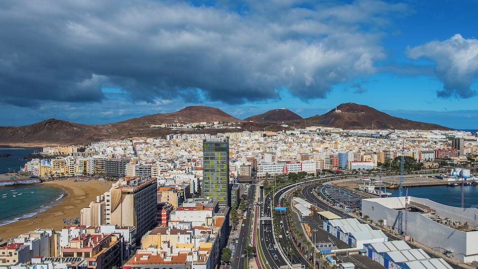 A coastal cityscape with a beach, mountains, and a dense urban area with buildings and roads.