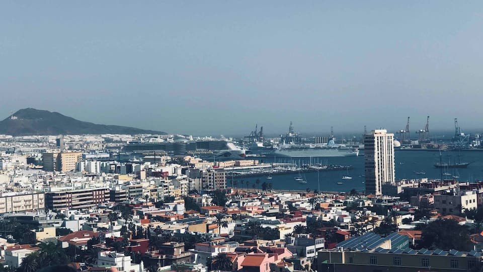 A coastal cityscape with a prominent cruise ship docked at the port, a large white building, and a mountain in the background.