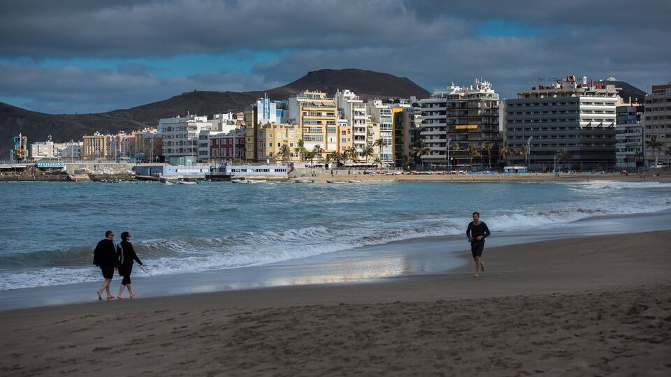 A beach with people walking, a cityscape in the background, and a mountain.