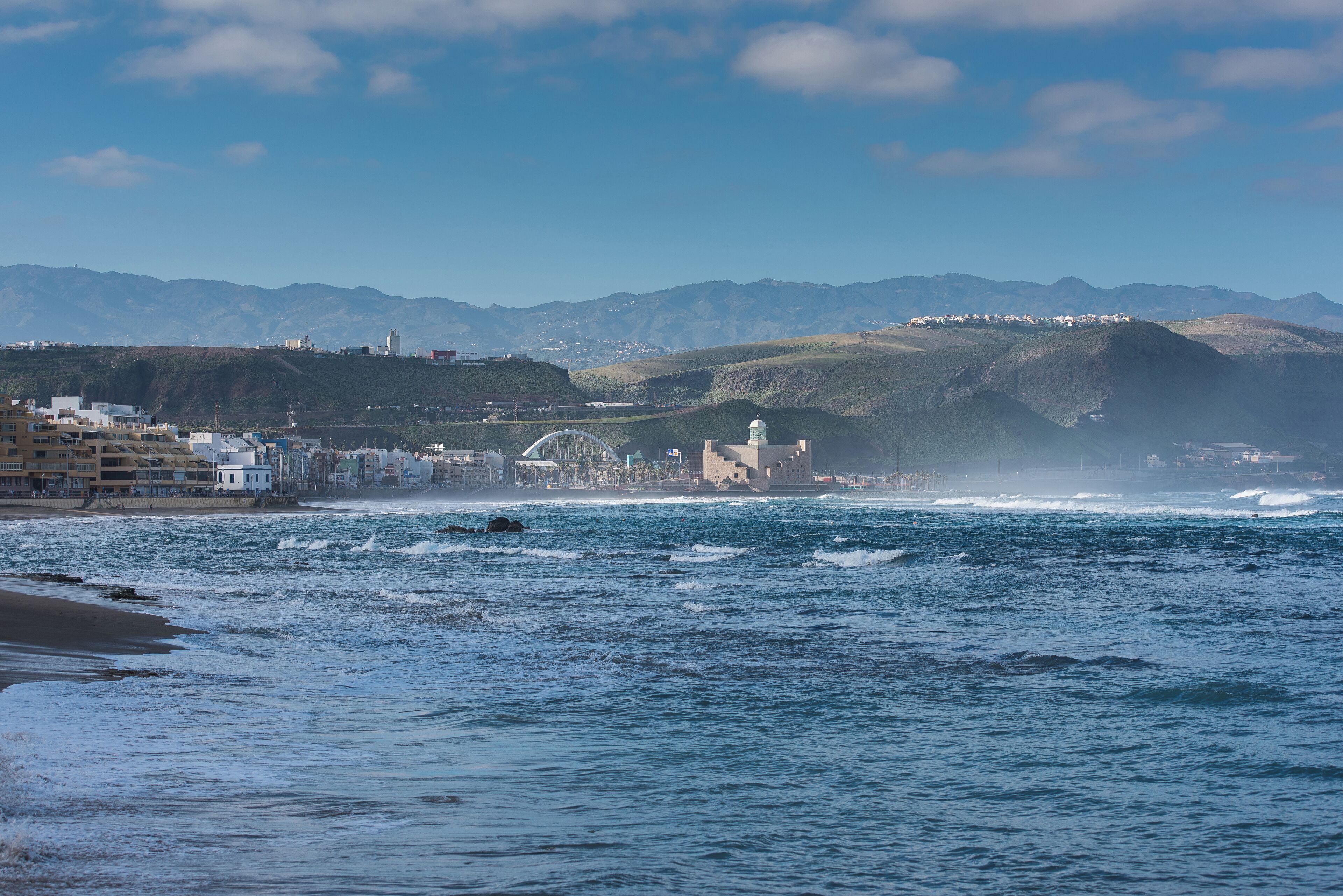 Un pueblo costero con una cúpula blanca destacada, una playa y montañas al fondo.