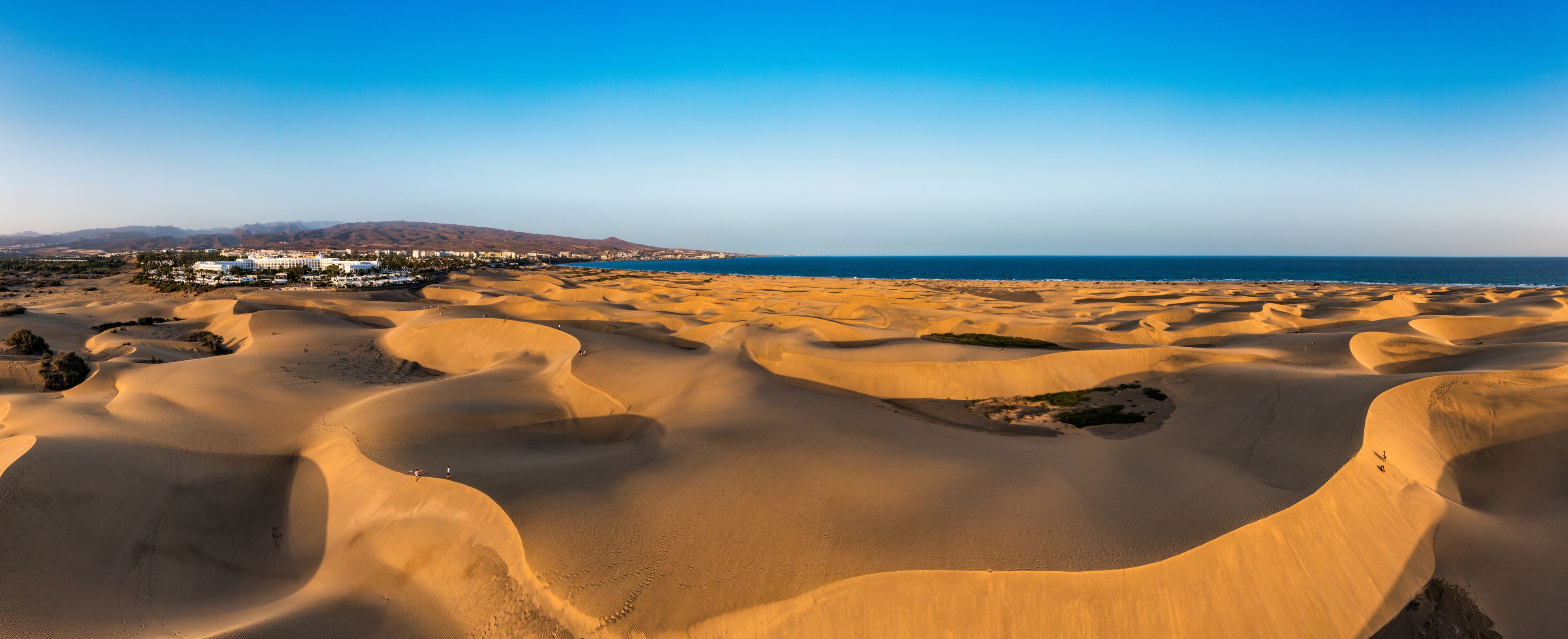 Paisagem desértica com dunas de areia, uma vila costeira e uma cordilheira ao longe.