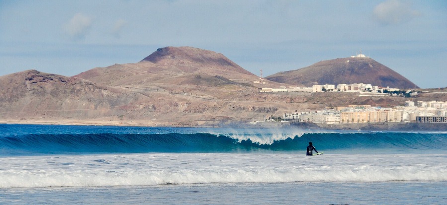 Un surfista sobre una ola, con montañas y un pueblo costero al fondo.