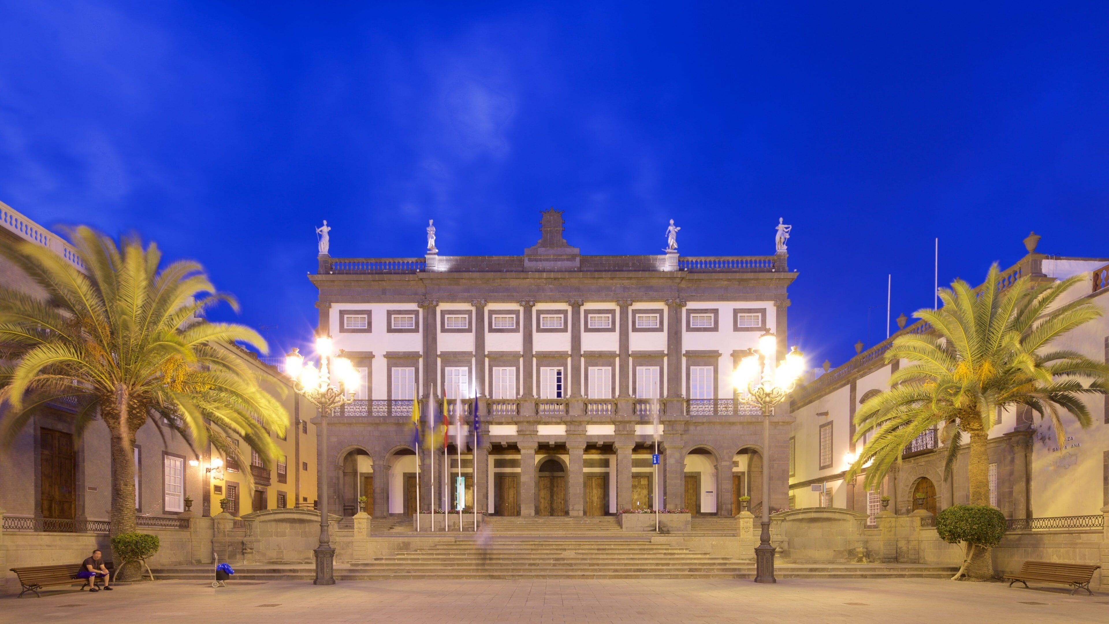A grand building with a central staircase, flanked by palm trees and illuminated by street lamps.
