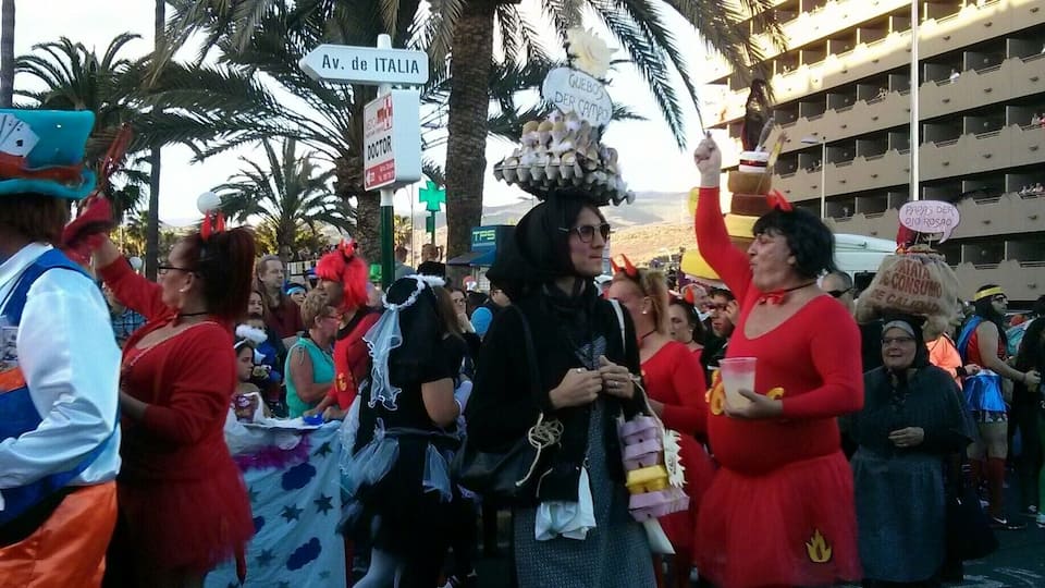 A parade with people in costumes, palm trees, and a building in the background.