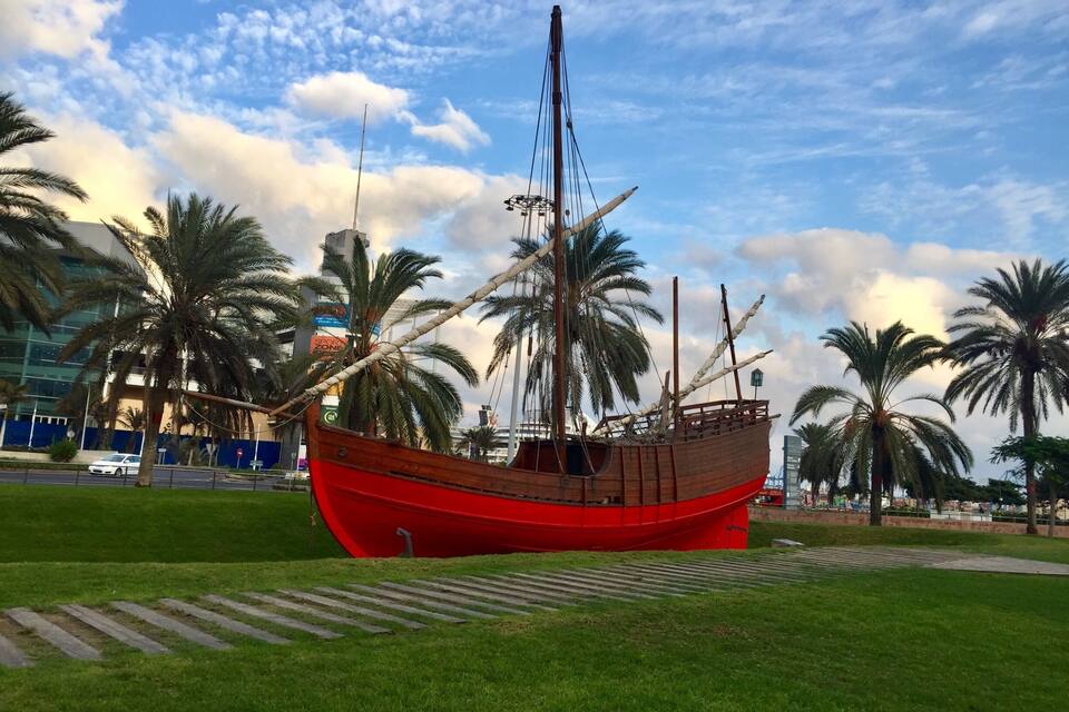 A red ship in a grassy area with palm trees and a modern building in the background.