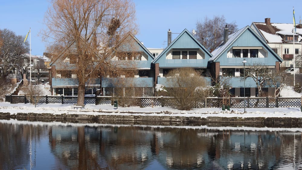 A row of blue houses by a calm river with snow on the ground.