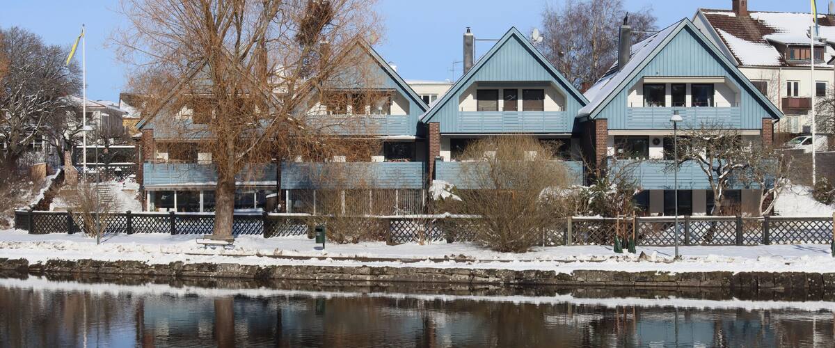 A row of blue houses by a calm river with snow on the ground.