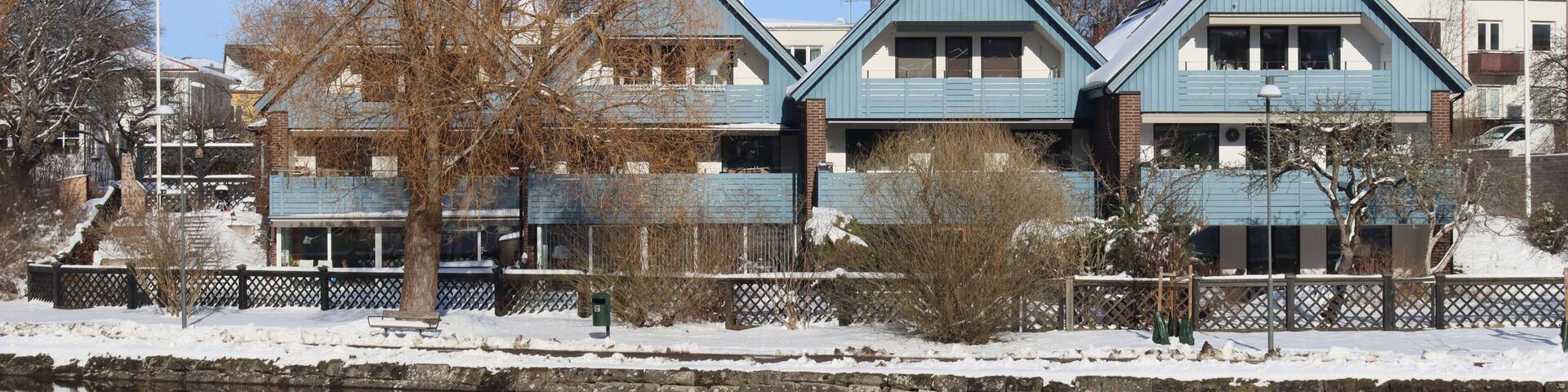 A row of blue houses by a calm river with snow on the ground.