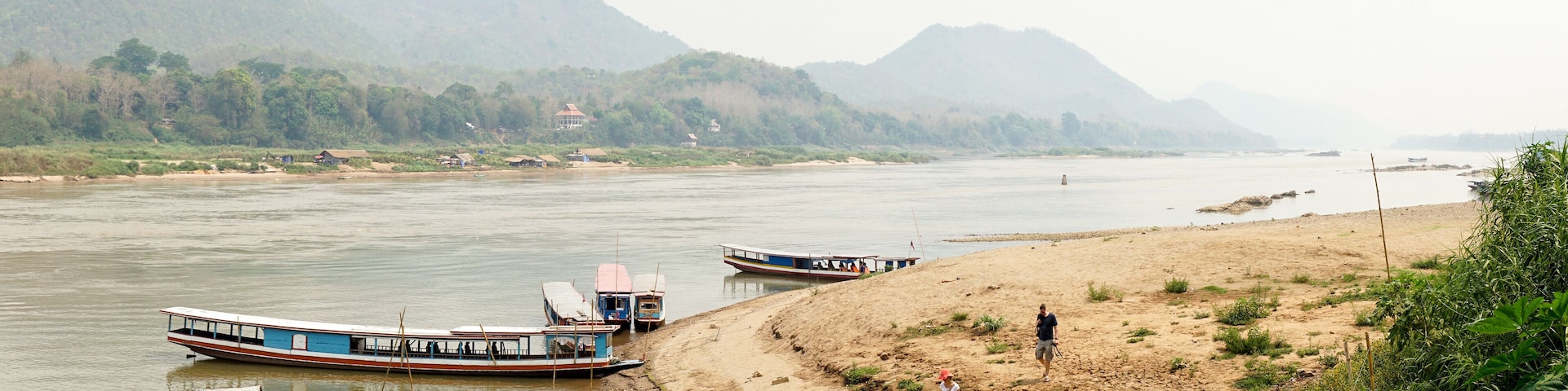 A river with boats, a sandy beach, and mountains in the background.