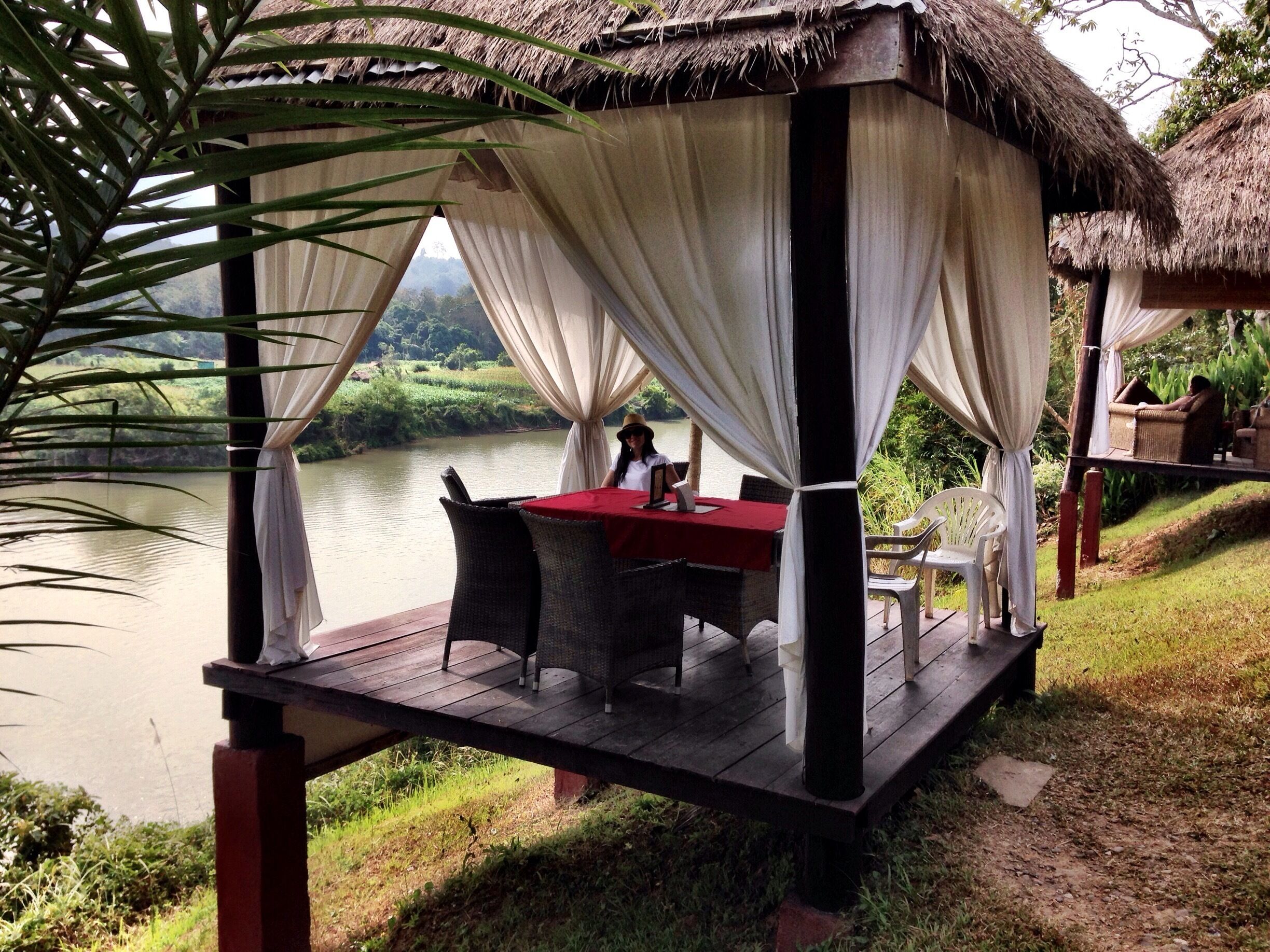 A thatched-roof gazebo with a table and chairs, overlooking a body of water.