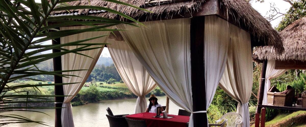 A thatched-roof gazebo with a table and chairs, overlooking a body of water.