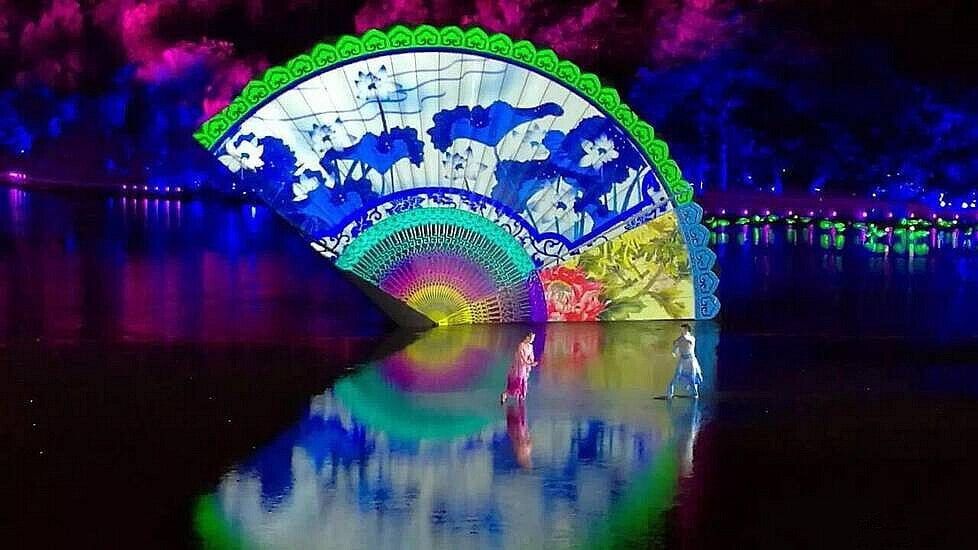 A large, illuminated fan structure with floral patterns reflecting on water.