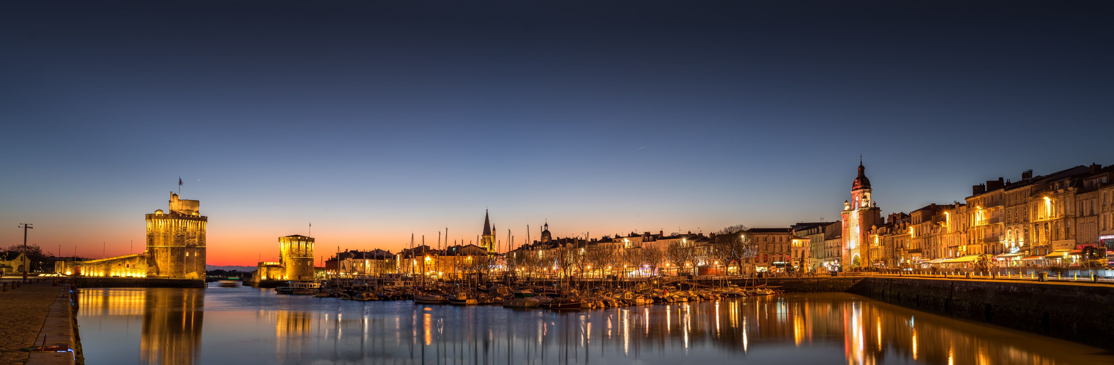 A cityscape at dusk with illuminated buildings, a calm river, and a prominent tower.