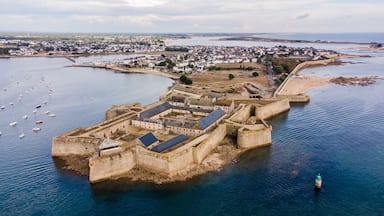 Una fortificación costera con una ciudad circundante, un puerto con barcos y una costa distante.