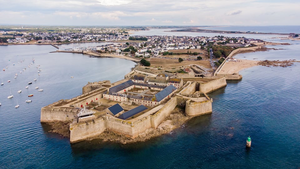 A coastal fortification with a surrounding town, a harbor with boats, and a distant shoreline.