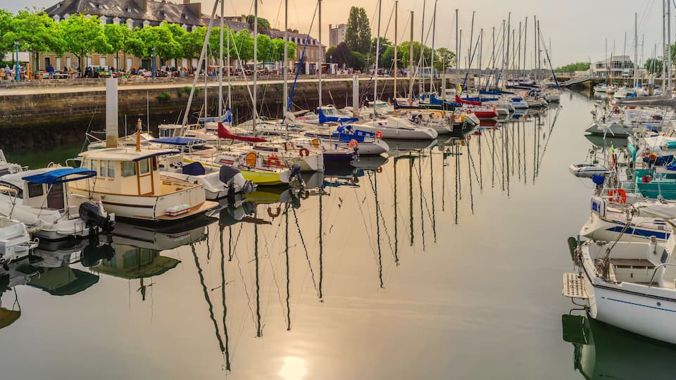 A marina with numerous boats docked along a calm waterway, surrounded by buildings and trees.